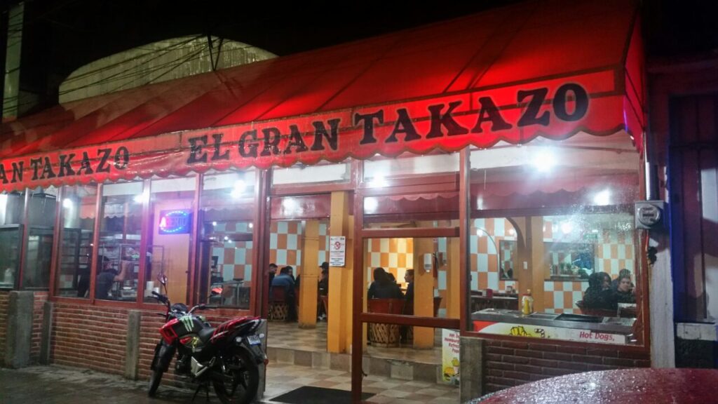 Nighttime entrance of El Gran Takazo restaurant in Puebla de Zaragoza with a motorcycle parked in front.